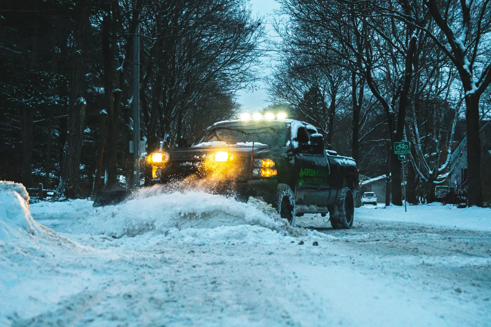 Greenworks plow truck pushing snow down a tree-lined road